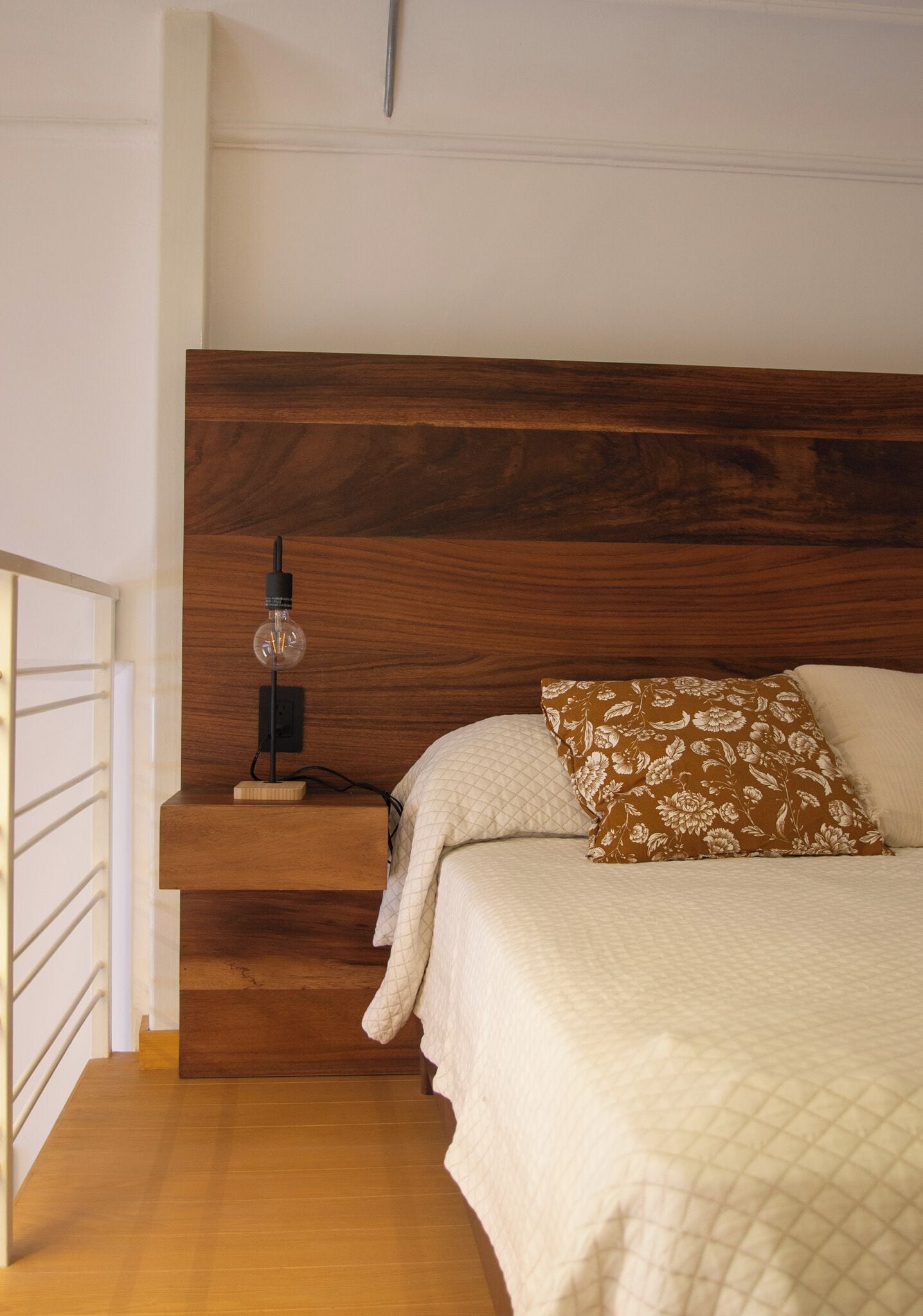 Un dormitorio con cabecera de madera, lámpara de noche, almohada estampada con flores y colcha acolchada.