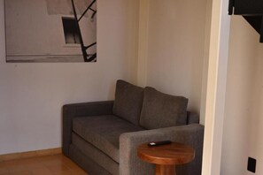 A corner of a room with a grey sofa, a small wooden side table, and a black remote control on the table. - Lopez Cotilla 658 (Guadalajara)
