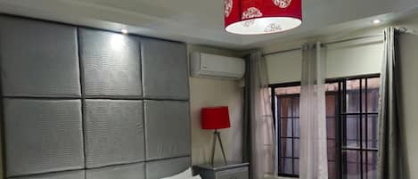 Una habitación de hotel con una cama grande, un cojín decorativo rojo, una lámpara roja sobre una mesita de noche y una ventana con cortinas.