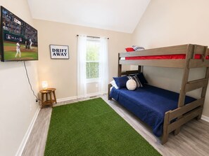 A bedroom with a bunk bed, a television mounted on the wall, a small table with a lamp, and a window with curtains.