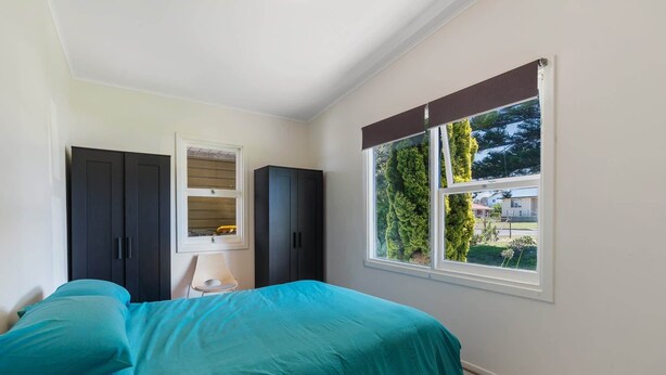 Un dormitorio con cama, dos armarios, una ventana que da a zonas verdes y una mesita con una silla.