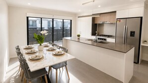 A modern kitchen with a central island, stainless steel appliances, and a dining area with a table set for six. - The Sunny Nook - 20 mins to BNE CBD (Coopers Plains)