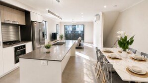 A modern kitchen with a central island, stainless steel appliances, and a dining area with a table set for four. - The Sunny Nook - 20 mins to BNE CBD (Coopers Plains)
