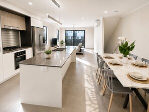 A modern kitchen with a central island, stainless steel appliances, and a dining area with a table set for four. - The Sunny Nook - 20 mins to BNE CBD (Coopers Plains)