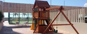 A wooden play structure with a swing set in a backyard with a view of palm trees and a building in the background. - Spacious 3BR Marina Condo Near Cancun Hotel Zone (Cancun)