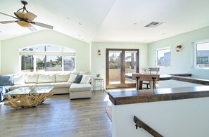 A spacious living room with a high ceiling, a ceiling fan, a glass coffee table, a sofa, and a dining area with a table and chairs. - Walk to Pierpont Sand · Private Rooftop Deck · Ocean Peek · 30+ Night Stays (Ventura)