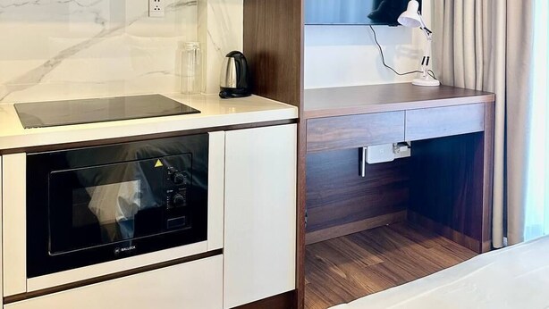 Una kitchenette moderna con horno integrado, microondas y cafetera. Hay un escritorio de madera con lámpara y un televisor de pantalla plana montado encima.