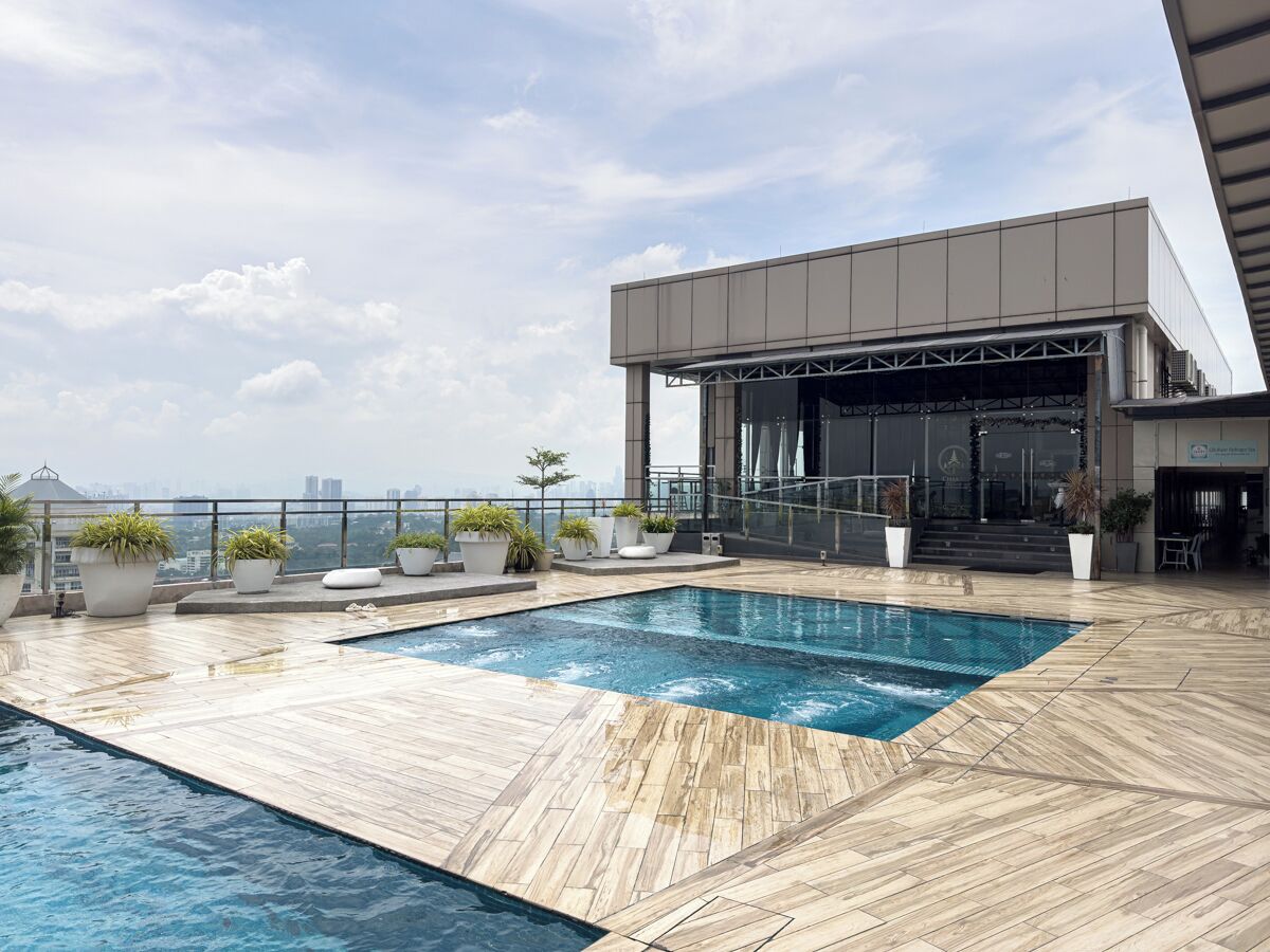 A rooftop pool with a wooden deck, surrounded by potted plants, and a modern building with a stage in the background.