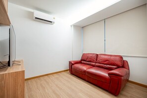 A room with a red leather sofa, a flat-screen TV on a wooden stand, and an air conditioning unit mounted on the wall. - Charoen One (Bangkok)