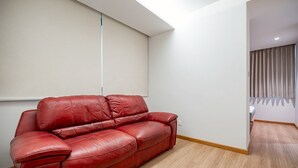 A red leather sofa in a room with light wooden flooring, white walls, and a partially visible bed in the background. - Charoen One (Bangkok)