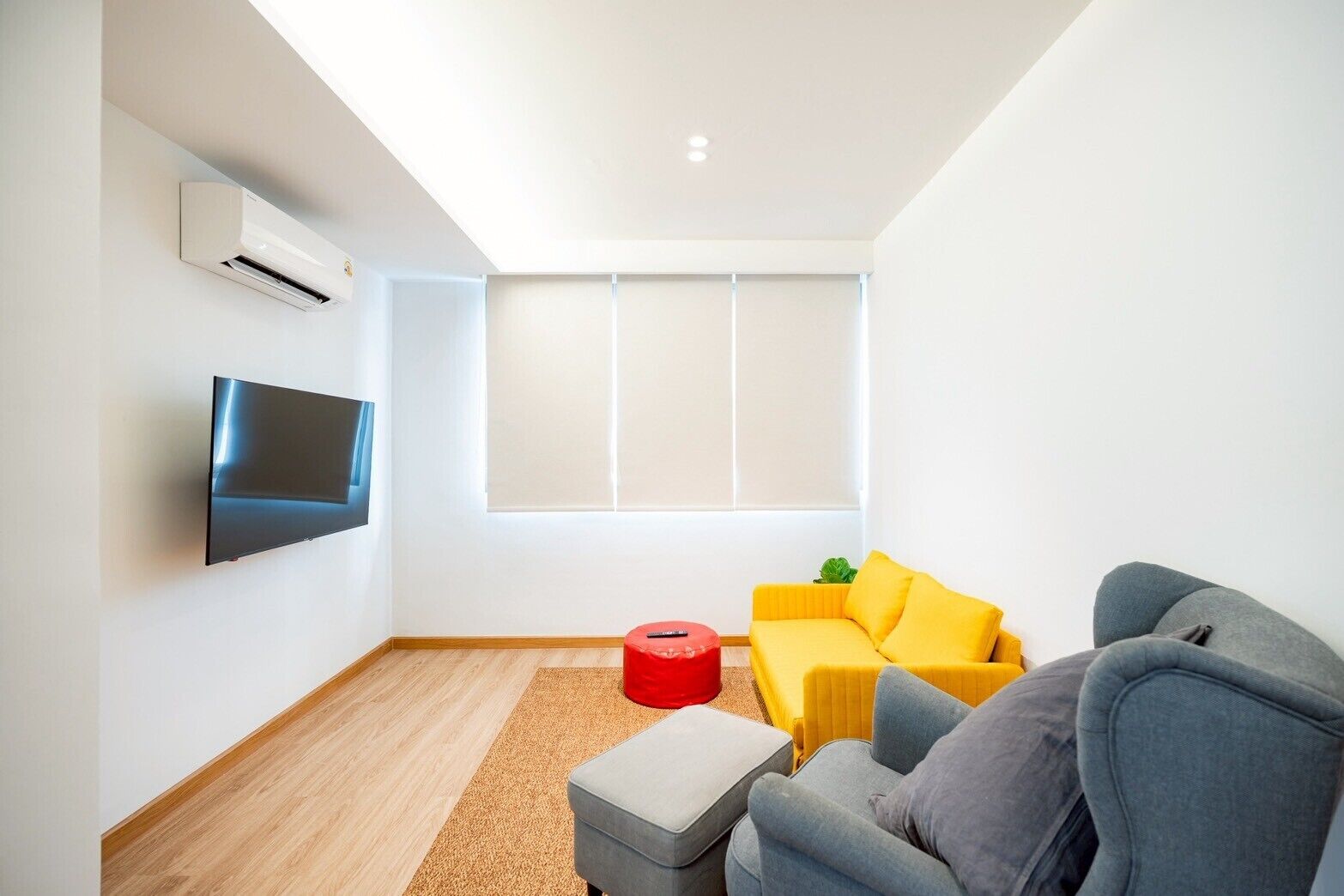 A modern living room with a flat-screen TV, a yellow sofa, a grey armchair, and a red ottoman.