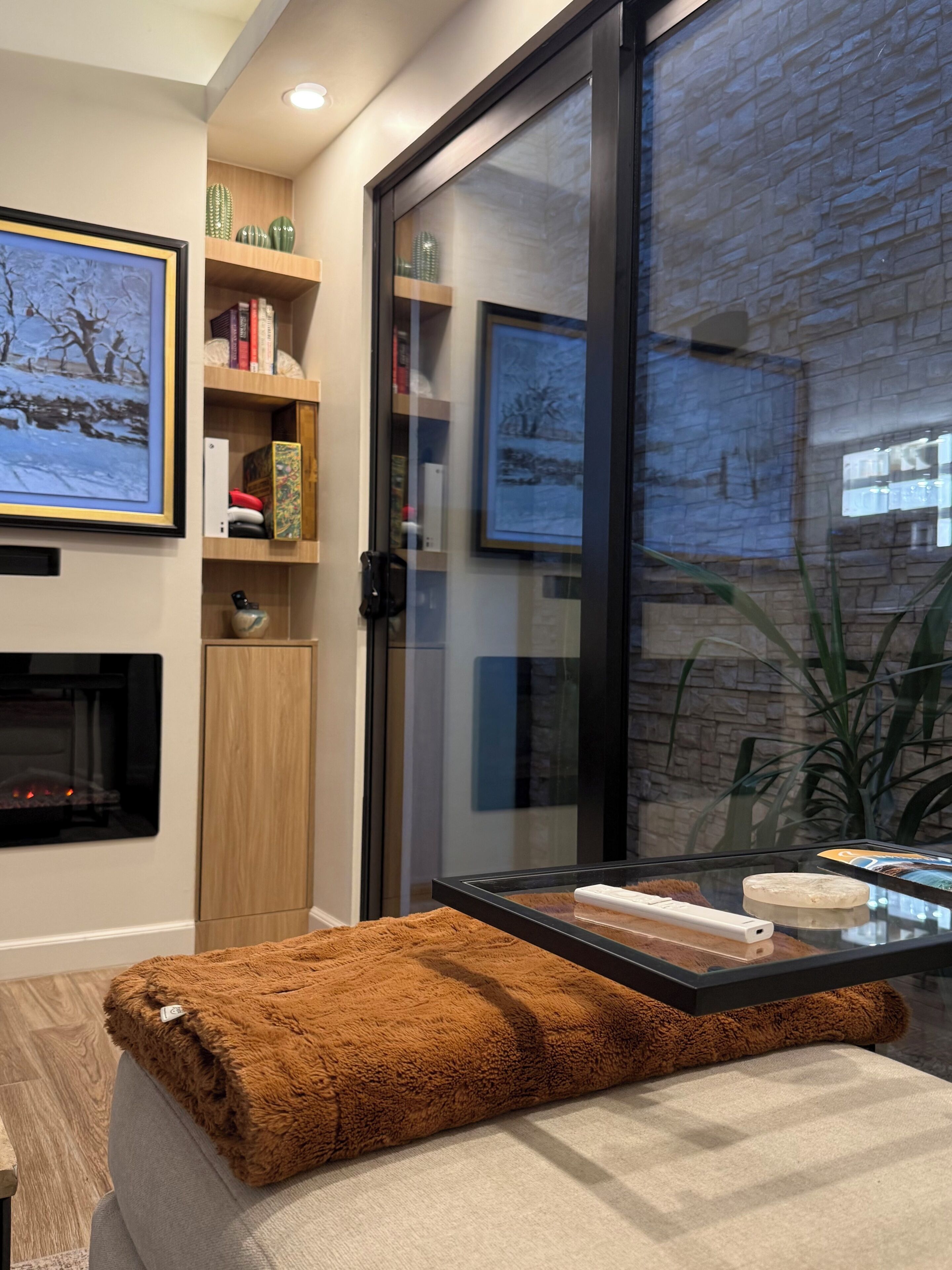 Uma sala de estar moderna com lareira, uma mesa de centro de vidro, um cobertor marrom e uma estante com diversos objetos.