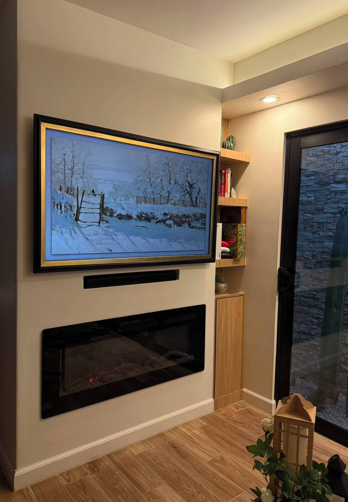 A living room with a flat-screen TV mounted on the wall, a fireplace below it, and a bookshelf to the right.