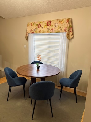 Ein Essbereich mit einem runden Holztisch, vier dunkelblauen Stühlen, einem Blumentopf und einem Fenster mit Jalousien und einem floralen Vorhang.
