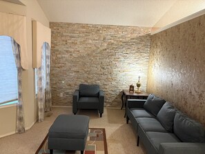 A living room with a stone wall, a grey sofa, a grey armchair, a small table with a lamp, and patterned curtains. - 
Holiday Home in West Edmonton (Edmonton)