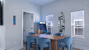 A room with a wooden table and four blue chairs, a gaming machine, and a surfboard decoration. - Hang Ten House- 2.5 Blocks to Beach- Pet Friendly (Myrtle Beach)