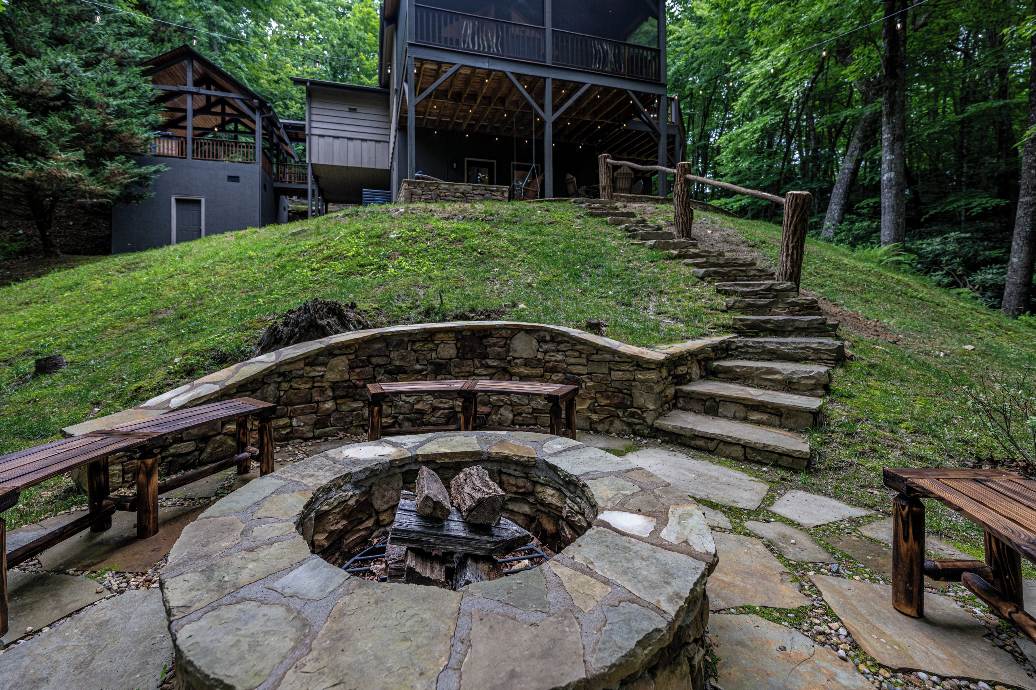 A stone fire pit with benches, a wooden staircase, and a building with a deck in a wooded area.