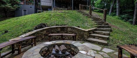 A stone fire pit with benches, a wooden staircase, and a building with a deck in a wooded area.