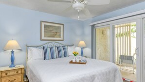 Ein Schlafzimmer mit einem Bett, zwei Nachttischlampen, einem Deckenventilator, einem gerahmten Bild und einer Glastür, die zu einem Außenbereich führt.