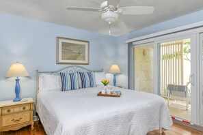 Ein Schlafzimmer mit einem Bett, zwei Nachttischlampen, einem Deckenventilator, einem gerahmten Bild und einer Glastür, die zu einem Außenbereich führt.