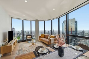 A modern living room with a city view, a flat-screen TV, a sofa, a coffee table, and a dining area set for two. - Live in the clouds! 3BD ultra-luxury in Southbank (Southbank)
