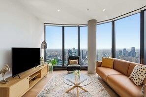 Ein modernes Wohnzimmer mit einem großen Flachbildfernseher, einer Couch, einem Couchtisch und Blick auf eine Stadtkulisse durch große Fenster.