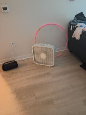 Una habitación con un ventilador de pie blanco, un aro de hula rosa y un objeto negro en el suelo.