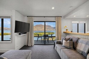 A modern living room with a flat-screen TV, a sofa, a dining area with chairs, and a large window offering a scenic view of mountains and a lake. - Jaw Dropping Views, Close To The Heart Of Queenstown (Queenstown)