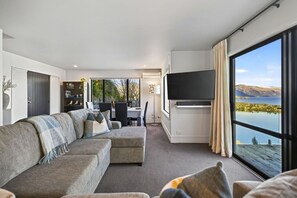 A modern living room with a sofa, a flat-screen TV, and a dining area with a table and chairs. - Jaw Dropping Views, Close To The Heart Of Queenstown (Queenstown)