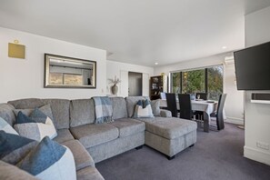 A modern living room with a sectional sofa, a dining area with a table and chairs, and a large flat-screen TV mounted on the wall.