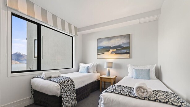 Quarto de hotel com duas camas, janela com vista para as montanhas, um quadro na parede e uma mesa pequena com abajur.
