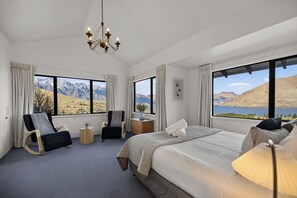 A hotel room with a large bed, two armchairs, a small side table, and a view of mountains and a lake.