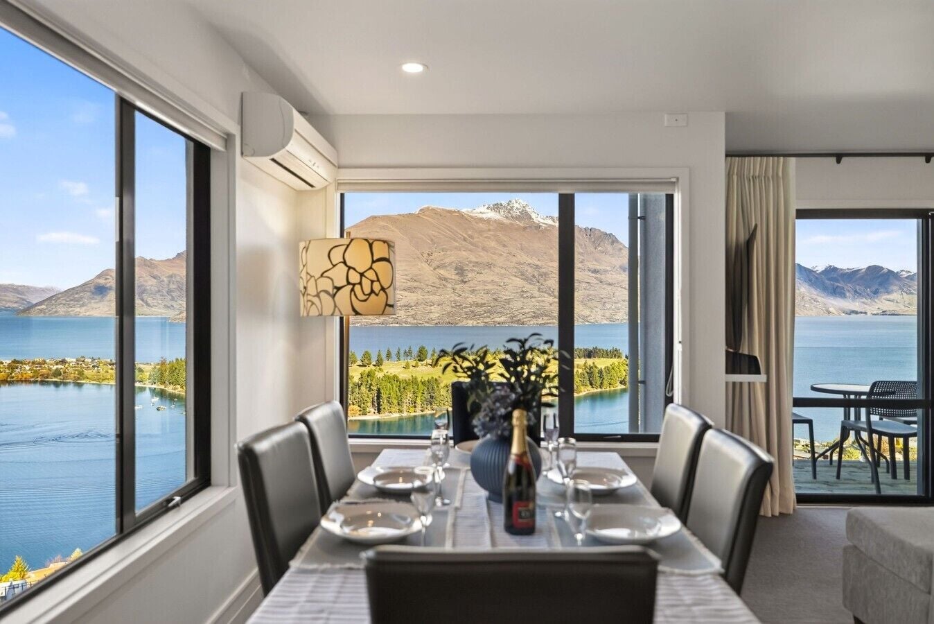 A dining area with a table set for four, large windows offering a view of a lake and mountains, and a modern air conditioning unit on the wall.