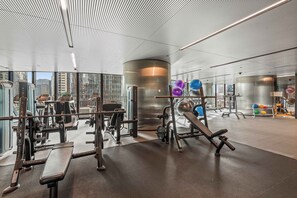 Ein modernes Fitnessstudio mit diversen Trainingsgeräten, einer großen zylindrischen Säule und Blick auf die Stadt durch große Fenster.