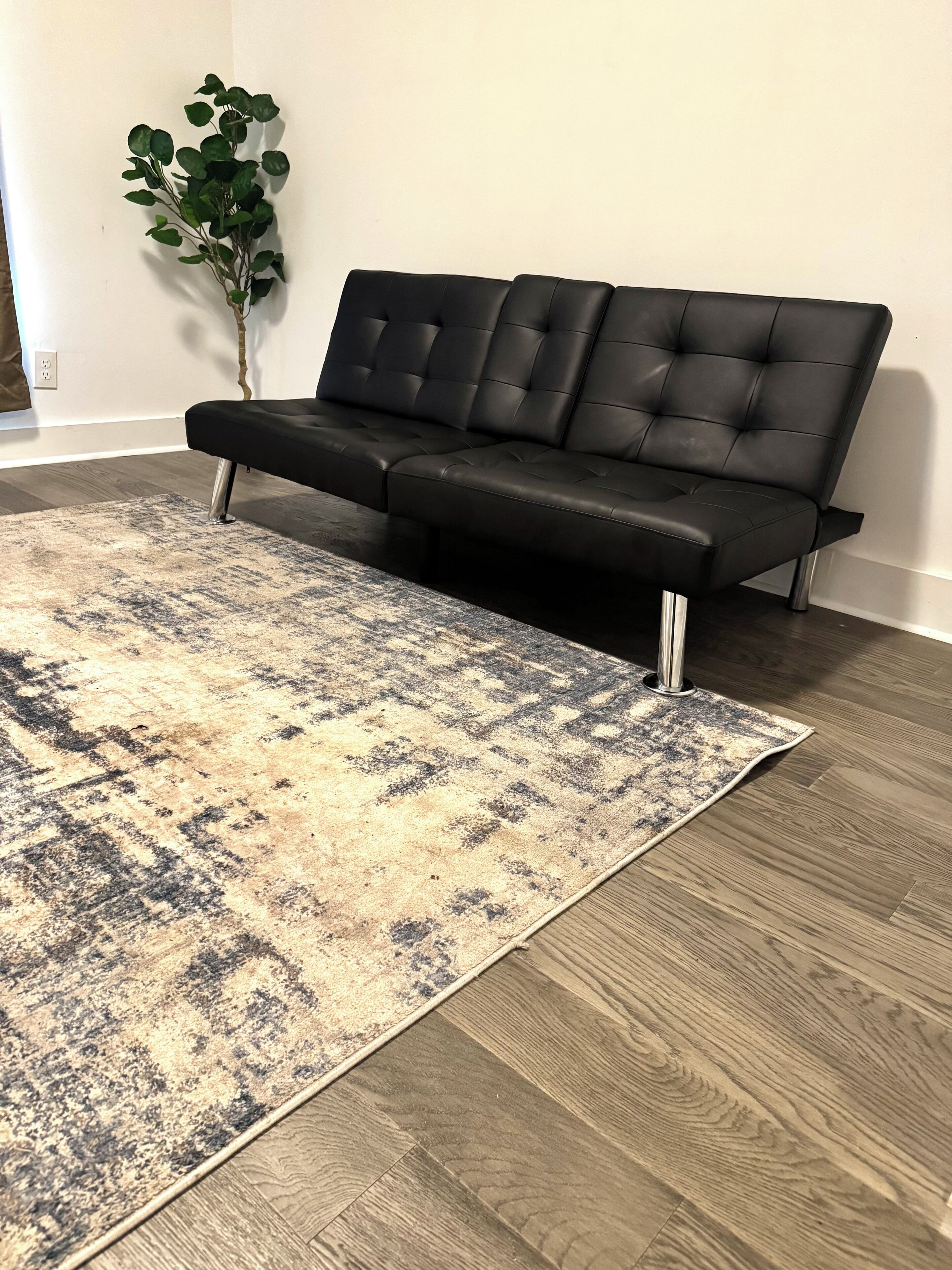 Un sofá de cuero negro con patas metálicas, una alfombra estampada y piso de madera.