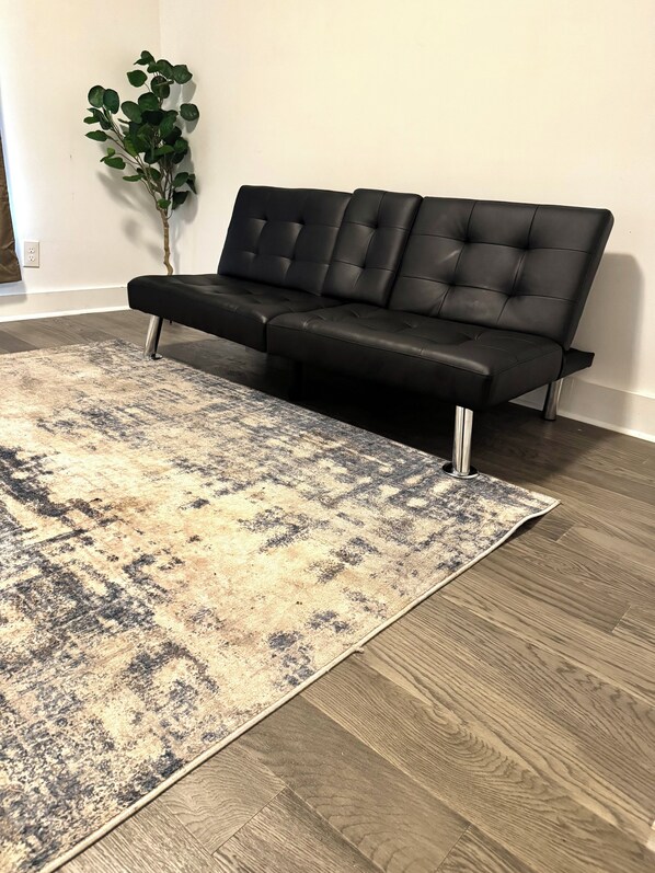 Un sofá de cuero negro con patas metálicas, una alfombra estampada y piso de madera.