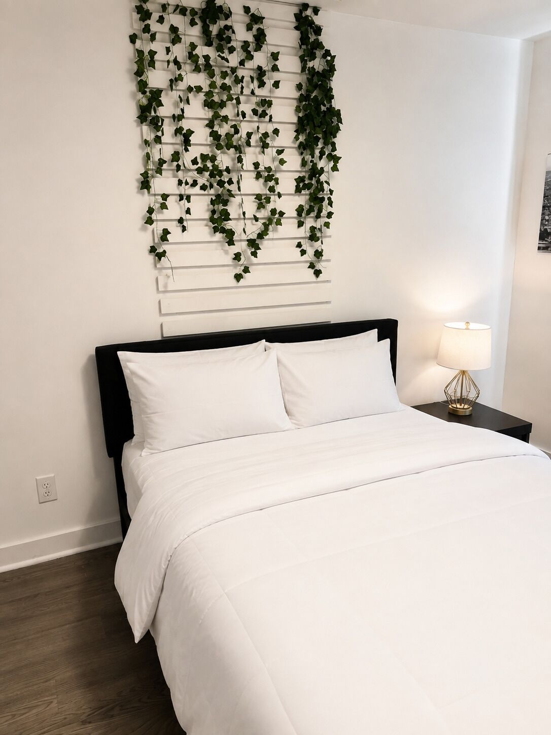 A bedroom with a bed, white bedding, a black headboard, a wall-mounted plant, a bedside lamp, and a wooden floor.