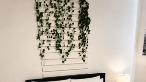 Un dormitorio con cama, ropa de cama blanca, cabecera negra, planta en la pared, lámpara de noche y suelo de madera.