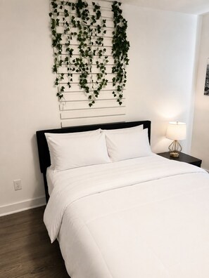 A bedroom with a bed, white bedding, a black headboard, a wall-mounted plant, a bedside lamp, and a wooden floor.