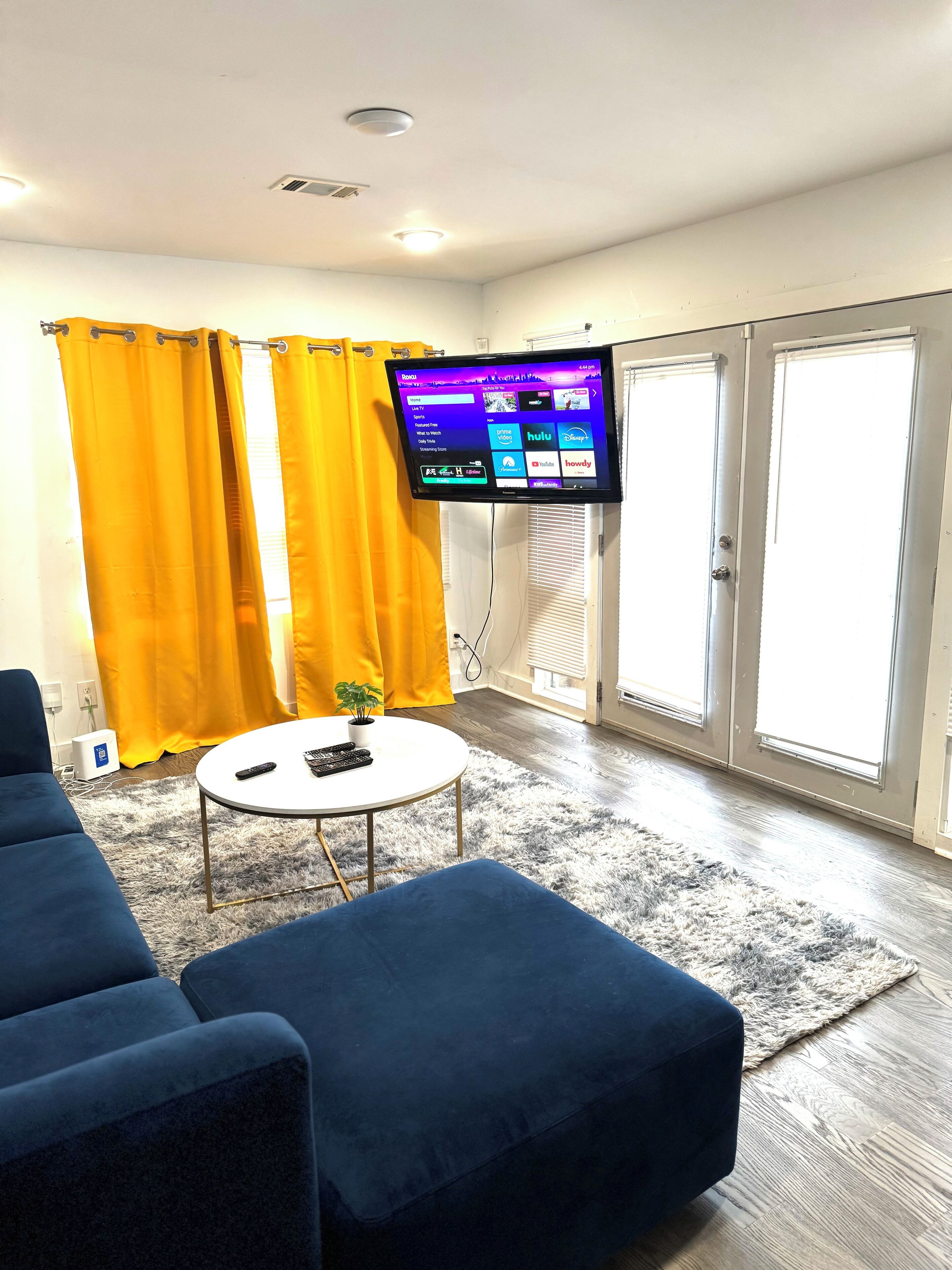 Un salon avec un téléviseur à écran plat fixé au mur, un canapé bleu, une petite table ronde avec une télécommande et une plante en pot.