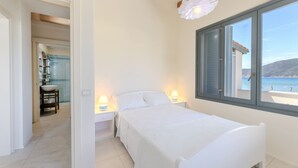A bedroom with a bed, bedside lamps, a window with a view of the sea, and a bathroom with a glass shower. - Villa Acquamarina - Gambero (Capoliveri)