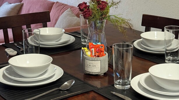 A dining table set with white plates, bowls, and cutlery, a vase with red roses, and a bottle with a label. - CM House with Parking (Hermosillo)