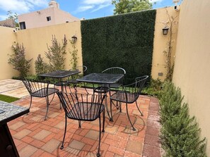 Une terrasse extérieure avec une table et des chaises, une haie verdoyante et un sol carrelé.
