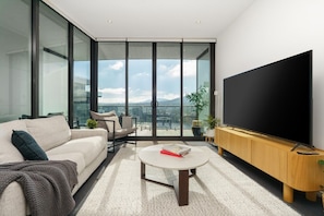 Ein modernes Wohnzimmer mit einem großen Flachbildfernseher, einer Couch, einem Couchtisch und Blick auf eine Stadtkulisse durch raumhohe Fenster.