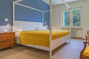 Un llit amb cortines, un edredó groc, una tauleta de nit de fusta, una làmpada penjada a la paret i una finestra amb vista a vegetació.