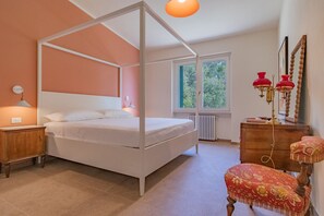 Ein Schlafzimmer mit einem Himmelbett, einer hölzernen Kommode, einem rot gepolsterten Sessel und einem Fenster mit Blick auf Grünflächen.
