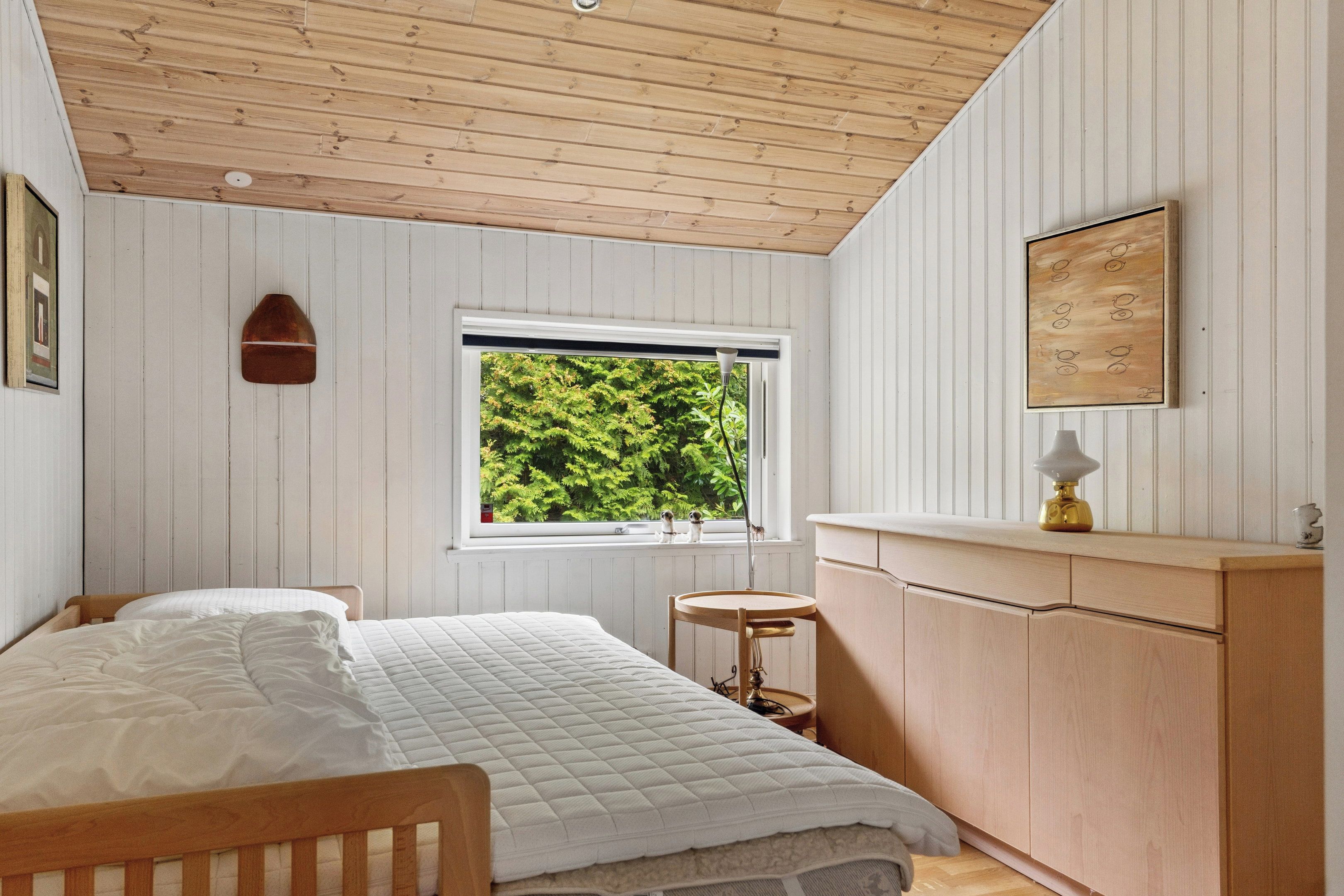 Une chambre avec un plafond en bois, une fenêtre donnant sur la verdure, un lit avec une literie blanche, un petit tabouret et une armoire en bois.