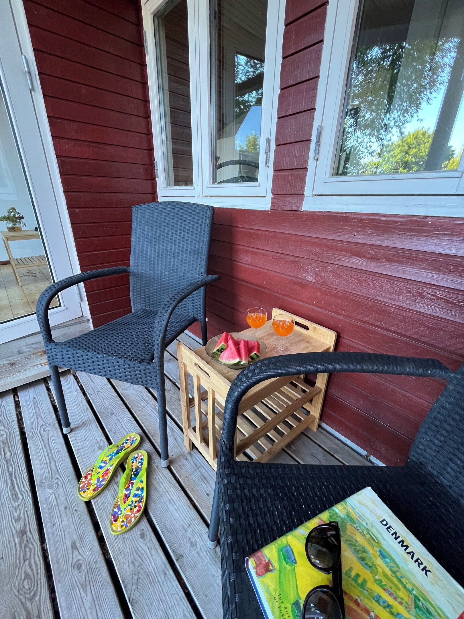A wooden deck with two wicker chairs, a wooden crate with a watermelon and drinks, and a book titled 'Denmark'.