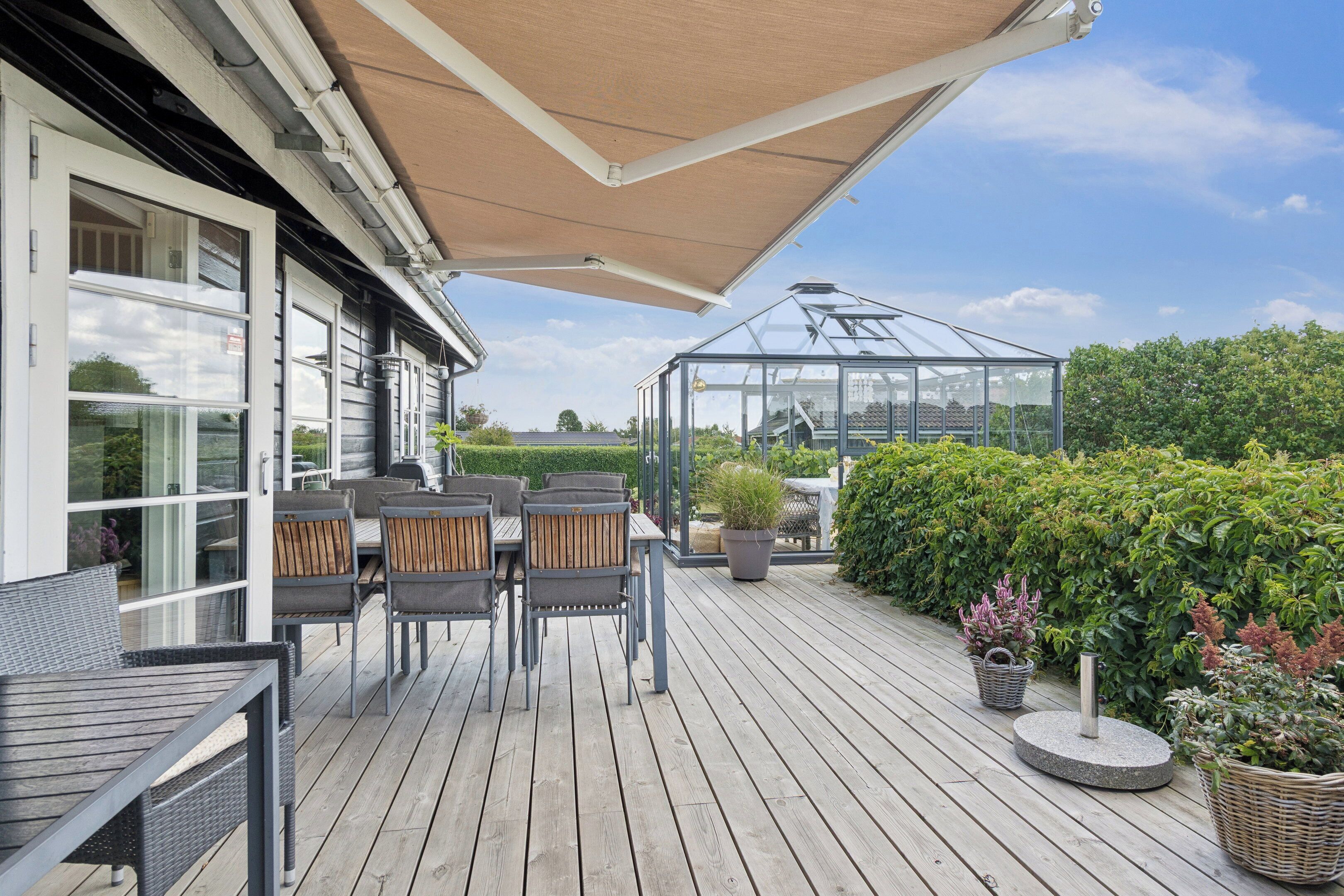 Une terrasse en bois avec une serre en verre, des meubles d’extérieur et une vue sur la verdure environnante.