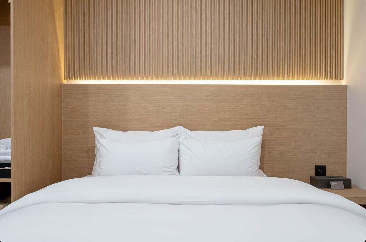 A modern hotel room with a wooden headboard, a neatly made bed with white linens, and a bedside table with a small electronic device.